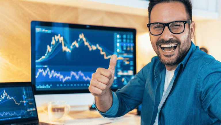 a cheerful man in a casual blue shirt and glasses, giving a thumbs-up gesture while sitting in front of multiple computer screens displaying financial charts. The charts feature upward and downward trends, suggesting active trading or financial analysis. The setting appears to be a modern, well-lit workspace, indicating a positive and successful trading experience, likely in cryptocurrency or stock markets. Tips for secure and controlling risk on crypto trading.